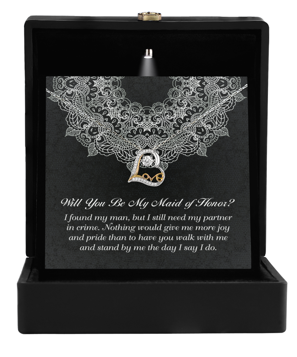 A black jewelry box is open, revealing the Love Dancing Necklace: a sterling silver piece adorned with a 14k gold and diamond-studded letter "L" pendant. Behind the necklace is a card asking, "Will You Be My Maid of Honor?" followed by a heartfelt message, all set against a lace-patterned background.