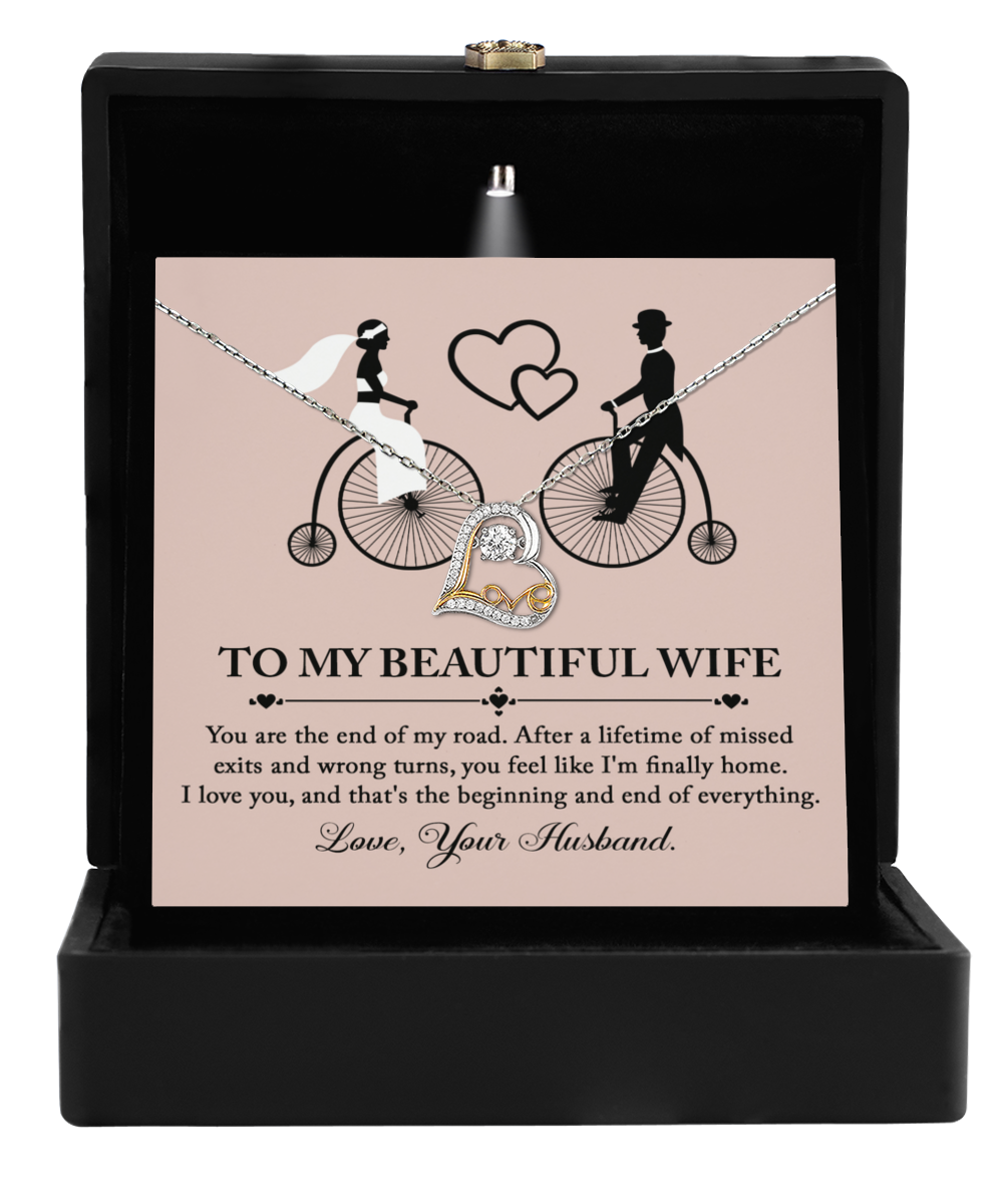 The "To Wife, I'm Finally Home - Love Dancing Necklace," showcasing intertwined rings, comes in a delightful gift box. Included is a card with the message "To My Beautiful Wife," accompanied by a romantic note and the silhouette of a couple on a tandem bike. It's the perfect gift for your wife.