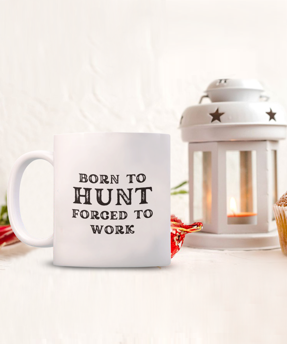 The "Born to Hunt Forced to Work" coffee mug, a limited-time offer, is set against a cozy scene with a candle lantern and cupcake. Printed and shipped from the USA, it's the perfect gift for hunters who cherish their passion.