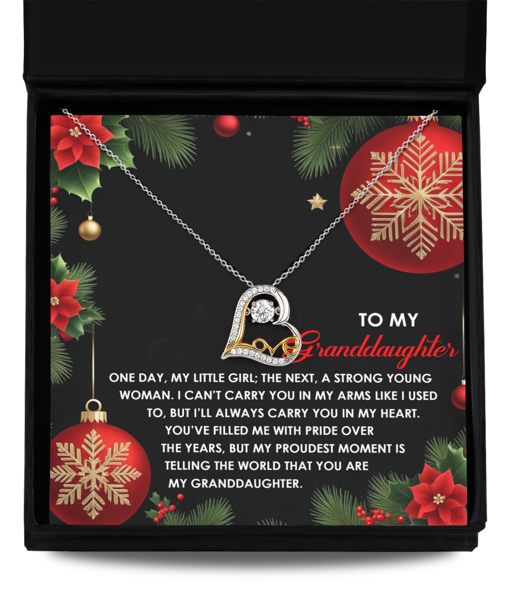 The Granddaughter-Proudest Moment - Love Dancing Necklace features a letter pendant nestled inside an open box, accompanied by a heartfelt message for a granddaughter. Surrounded by festive red and gold decorations against a holiday-themed backdrop, this 14k gold piece exudes warmth and affection.