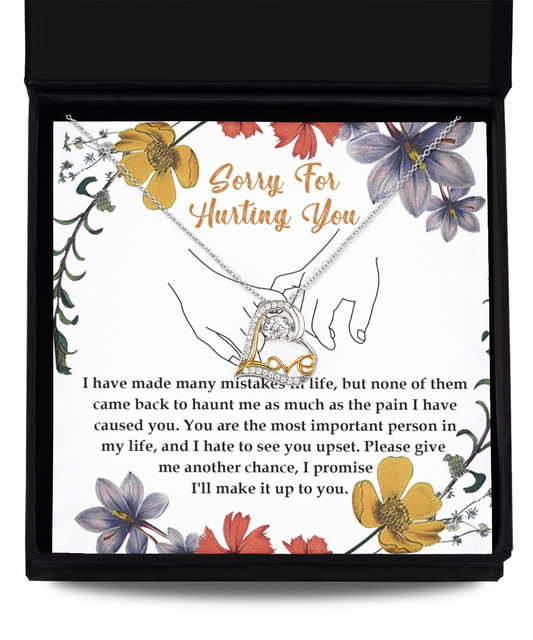 A stunning Sorry-Haunt Me - Love Dancing Necklace featuring an intertwined heart pendant, crafted in sterling silver, rests on a card that reads, "Sorry for hurting you," along with an apology message below. The card is beautifully decorated with floral illustrations.