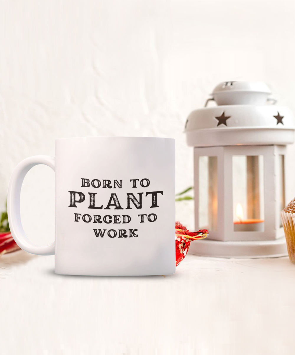 Next to a white lantern holding a candle and a muffin, the "Born to Plant Forced to Work" coffee mug is an exclusive design available only for a limited time, printed and shipped from the USA. Perfect gift idea for gardeners!.