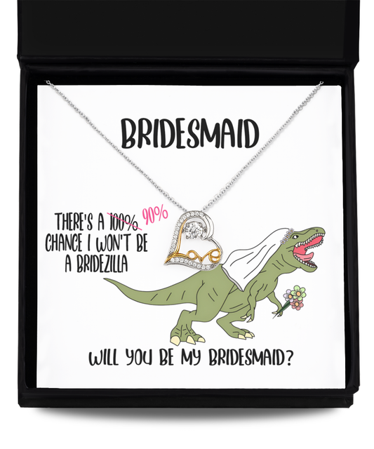 A Bridesmaid-Will You Be - Love Dancing Necklace is displayed on a card inside a black box. The card features a cartoon dinosaur in a veil holding flowers, with text asking, “Will you be my bridesmaid?”.