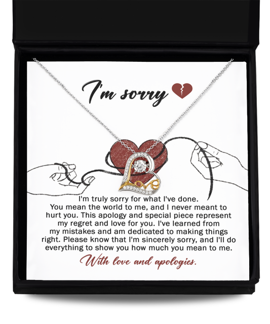 The "Sorry-Mean The World - Love Dancing Necklace," featuring a heart pendant, is displayed in a black box. The interior of the box is lined with 14k gold and includes an apology message expressing regret and love, along with an illustration of two hands holding red string.