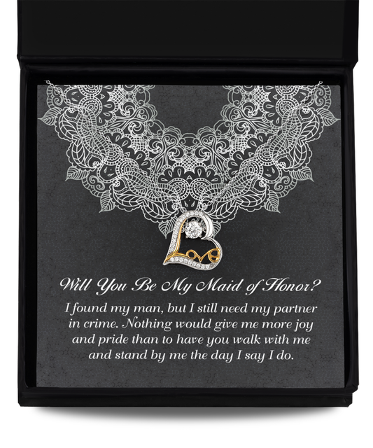 The "To Maid Of Honor-Partner In Crime - Love Dancing Necklace" comes in a box that contains a rhodium-plated necklace featuring a heart-shaped pendant with an embedded gem. The interior of the box has a lacy white design with the text: "Will You Be My Maid of Honor?" and below it reads: "I found my man, but I still need my partner in crime...".