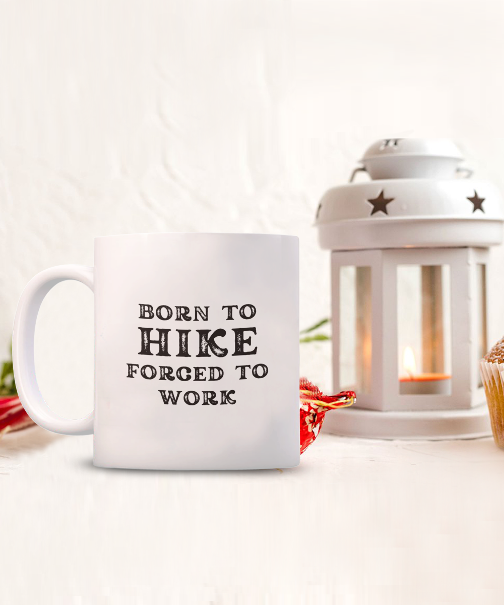 The "Born to Hike Forced to Work" Coffee Mug, ideal as a gift for hikers, is shown next to a lit white lantern candle. Available for a limited time; printed and shipped from the USA.