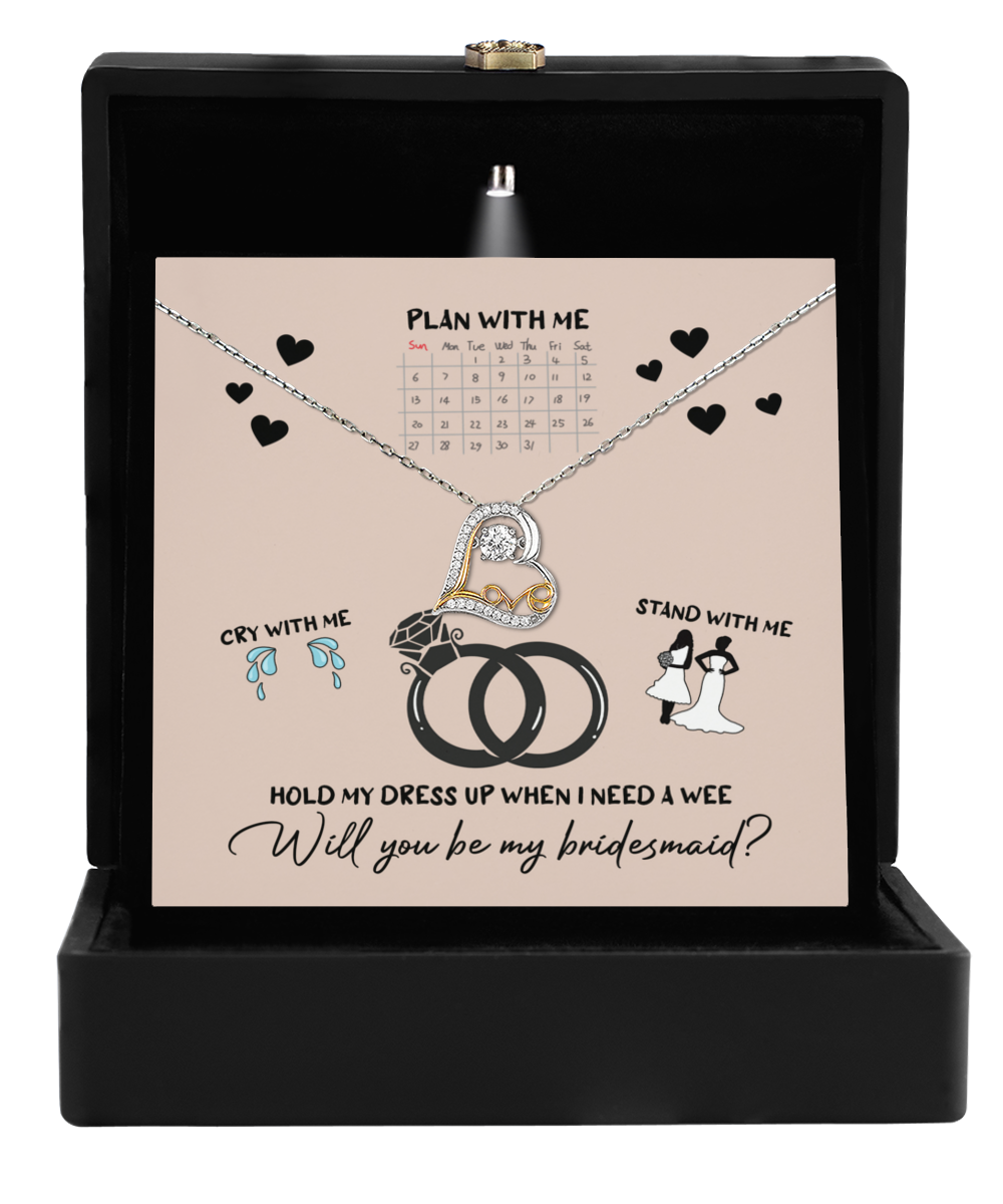 An open jewelry box in black reveals an elegant design, showcasing a card that features the Love Dancing Necklace. The card sweetly asks, "Will you be my bridesmaid?" and is adorned with charming graphics of rings, a calendar, and small figures—making it an unforgettable bridesmaid gift.