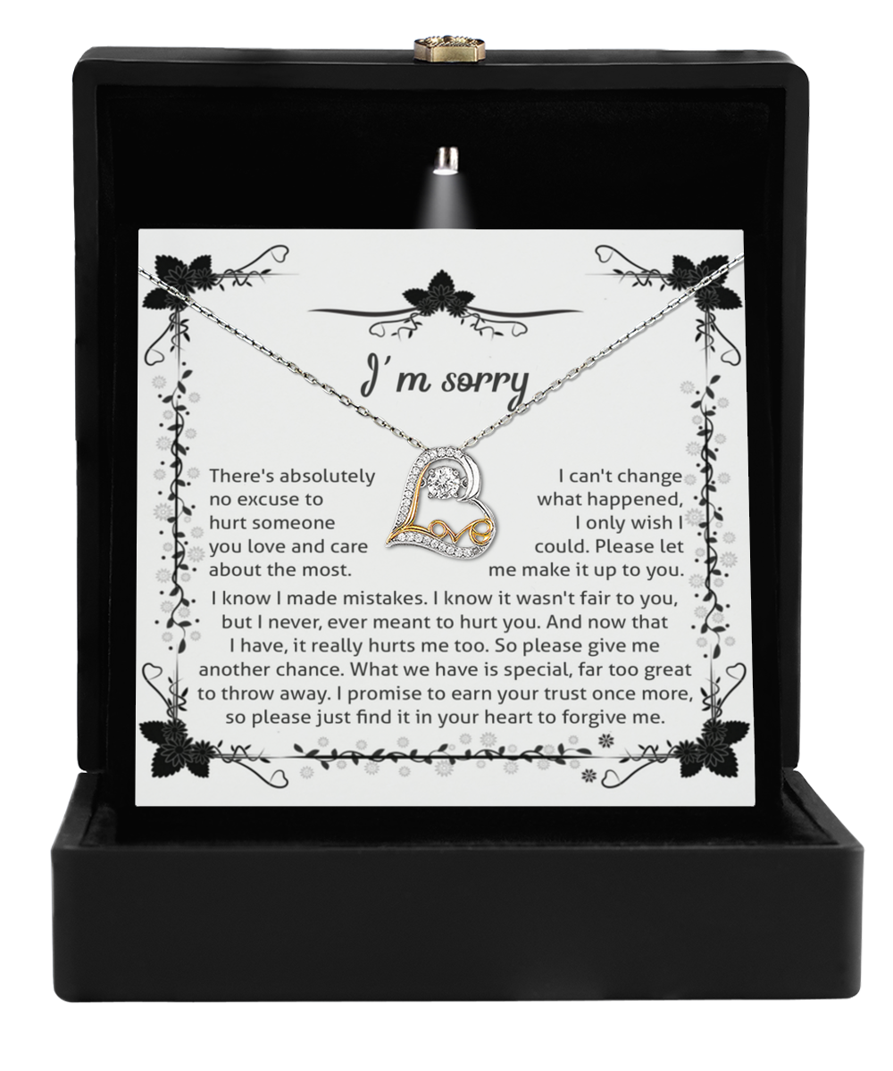 A Sorry-No Excuse - Love Dancing Necklace, featuring a silver heart shape accented with 14k gold and small crystals, is displayed in an open black box. Behind the necklace, a card reads “I’m sorry” with an apologetic message surrounded by black floral designs.