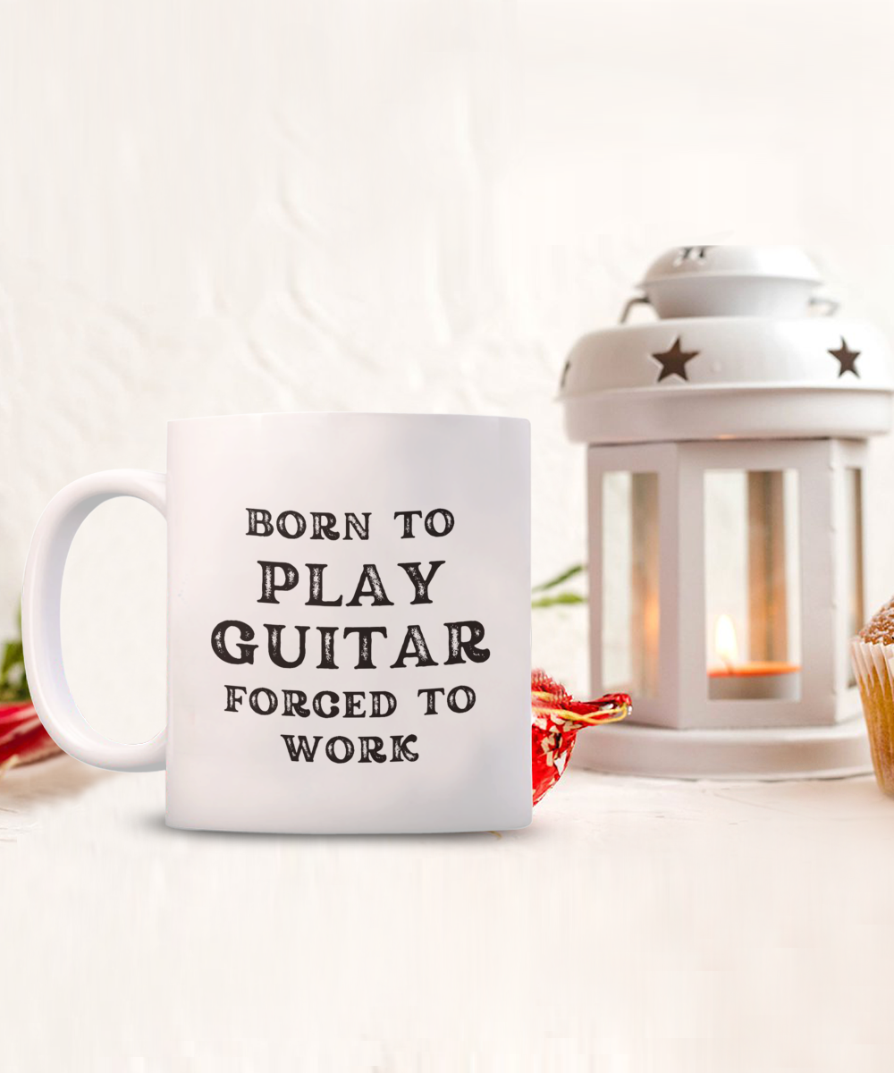 The "Born to Play Guitar Forced to Work" mug, a perfect gift for guitarists, is accompanied by a white lantern with a lit candle, red flower, and cupcake. This charming scene is printed and shipped from the USA and available for a limited time only.