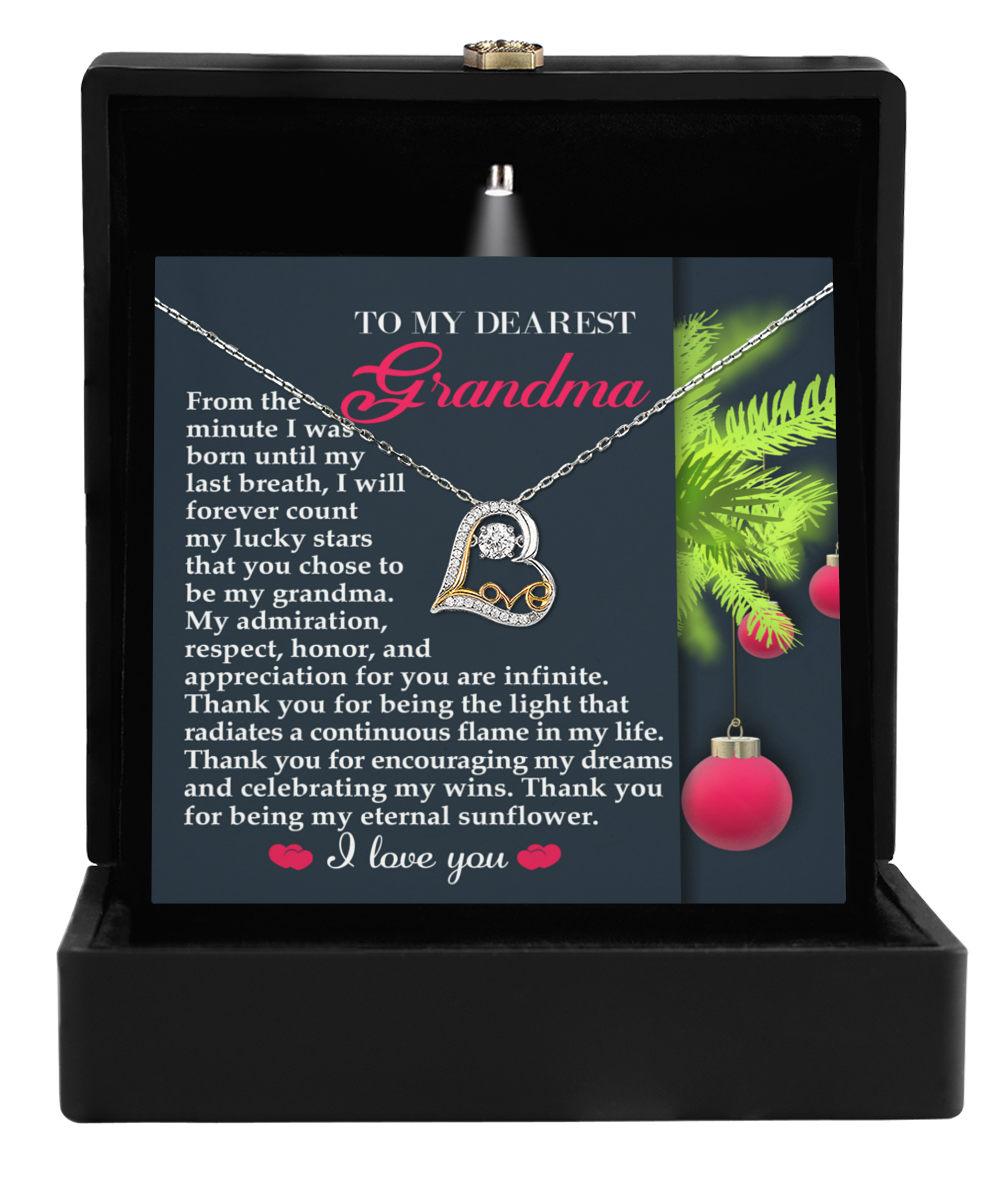 The Grandma-Eternal Sunflower - Love Dancing Necklace, adorned with a heart pendant, is elegantly displayed in a black box. Behind it, a touching Christmas-themed message celebrating maternal love and "Grandma" shines amid festive branches and ornaments.