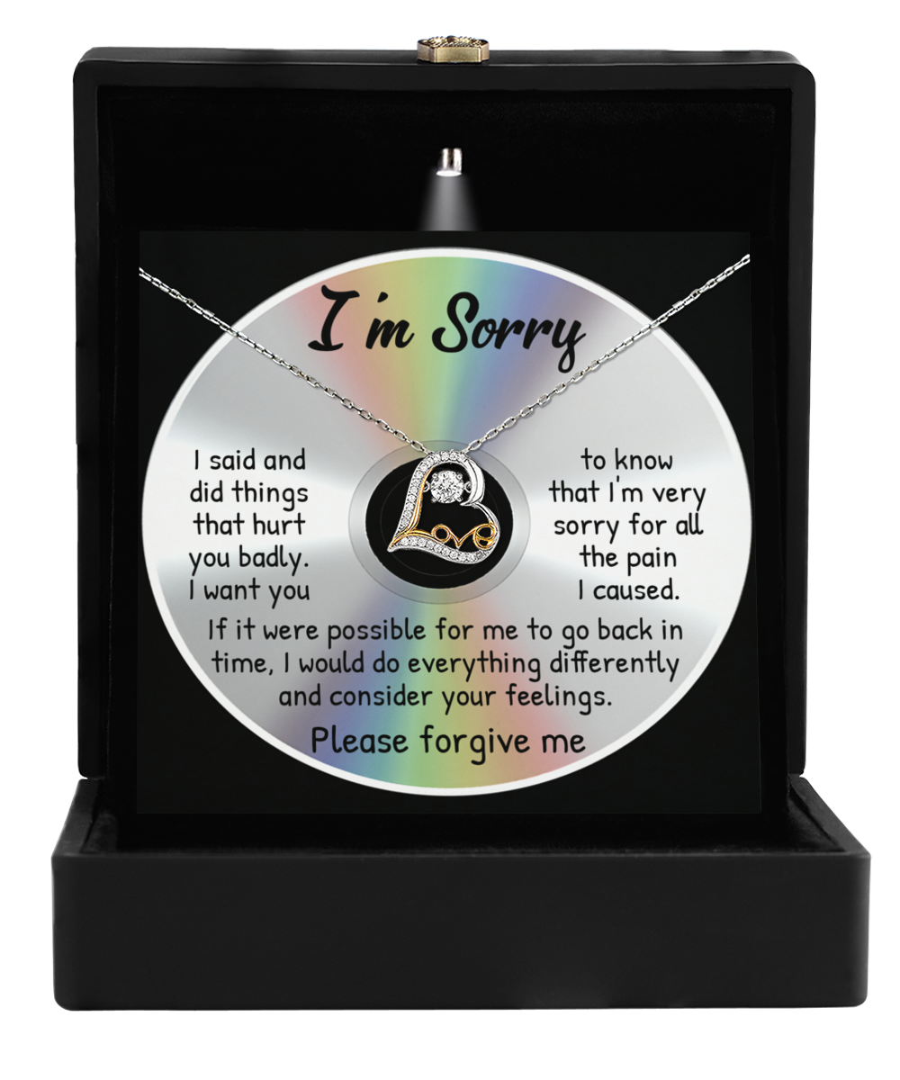 The Sorry-Back In Time - Love Dancing Necklace, featuring a heart pendant in sterling silver, is elegantly displayed inside an open black box. Behind the necklace is a disc with a printed apology message.