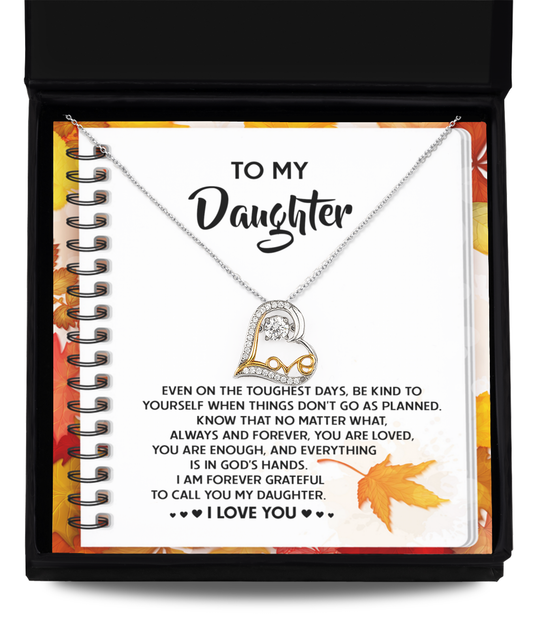 A Daughter-In God's Hands - Love Dancing Necklace, crafted in sterling silver and shaped as a heart, rests on an open notebook with a message beginning "To My Daughter" and inspiring words about love and resilience. Autumn leaves adorn the background, enhancing the sentimental charm of this touching scene.