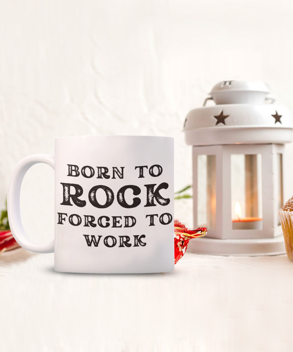 A "Born to Rock Forced to Work" white coffee mug sits beside a lit lantern and cupcake. This exclusive piece is not available in stores, available for a Limited Time Only, and printed and shipped from the USA. Ideal gift or appreciation cup for rock enthusiasts!.