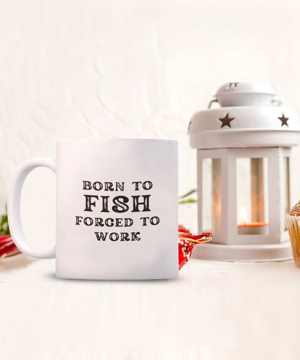 A "Born to Fish Forced to Work" coffee mug from the USA sits beside a lit lantern and a muffin on a white backdrop. This quirky angler gift is only available for a limited time.