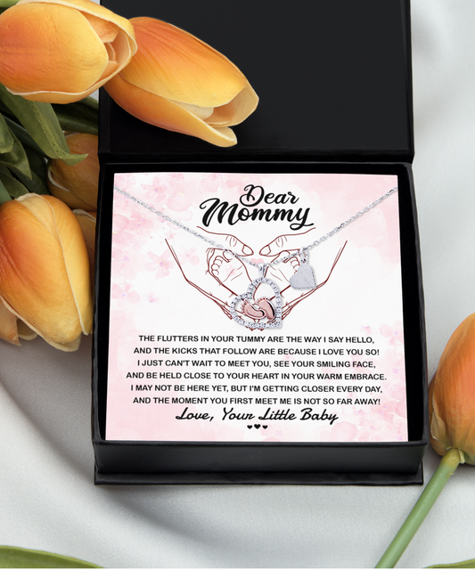 A close-up image of the "To Mommy, I Love You" - Baby Feet Necklace in a black gift box with a touching message from an unborn baby, surrounded by orange tulips.