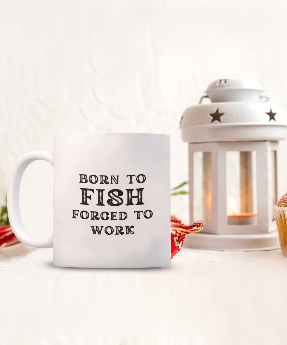 The "Born to Fish Forced to Work Coffee Mug" sits next to a lit candle lantern and a cupcake on a light surface. This delightful mug, printed and shipped from the USA, is available for a limited time only.