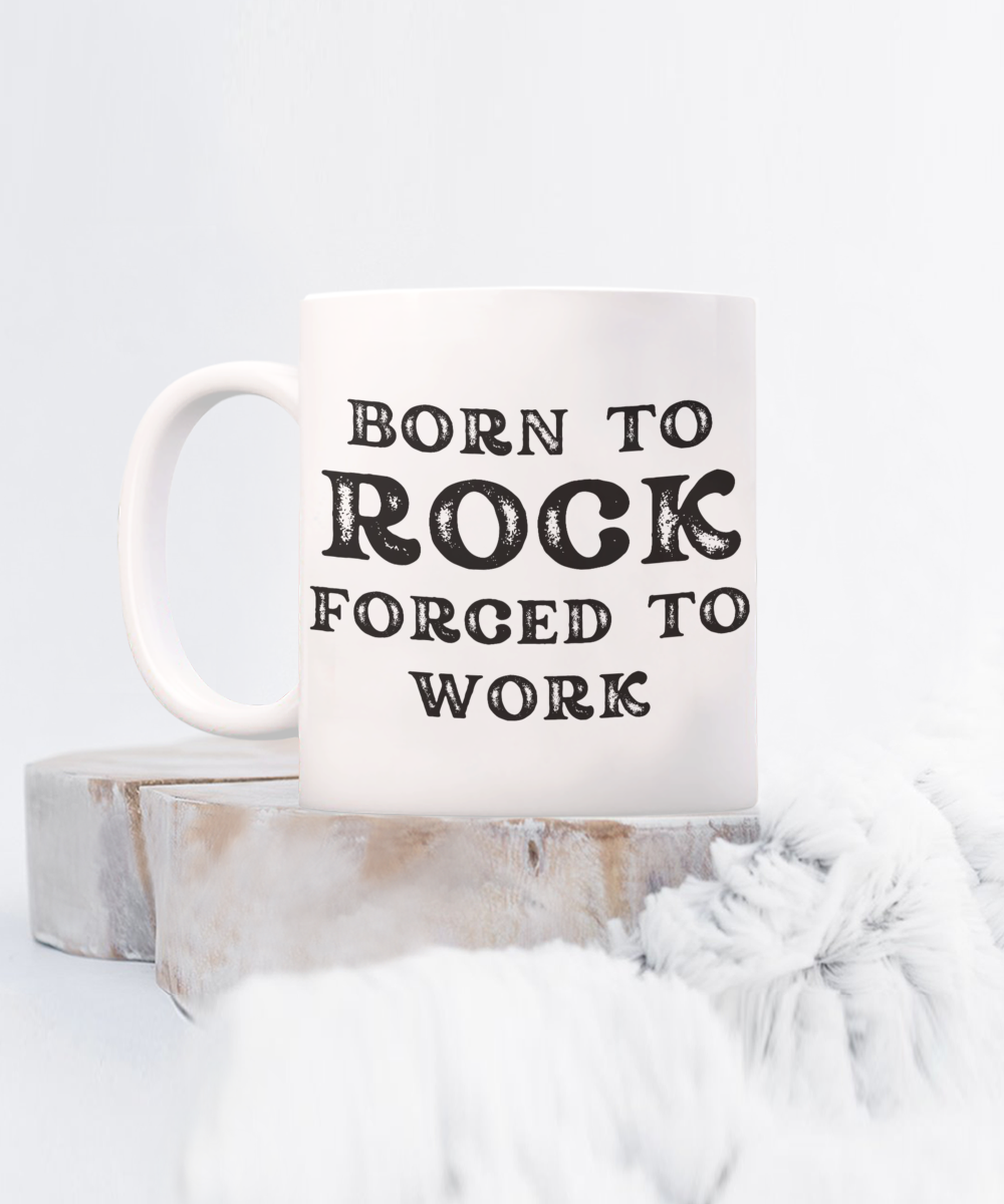 The "Born to Rock Forced to Work" coffee mug, featuring an exclusive design, is set on wooden slices against a fluffy white background. Printed and shipped from the USA, it's available for a limited time.