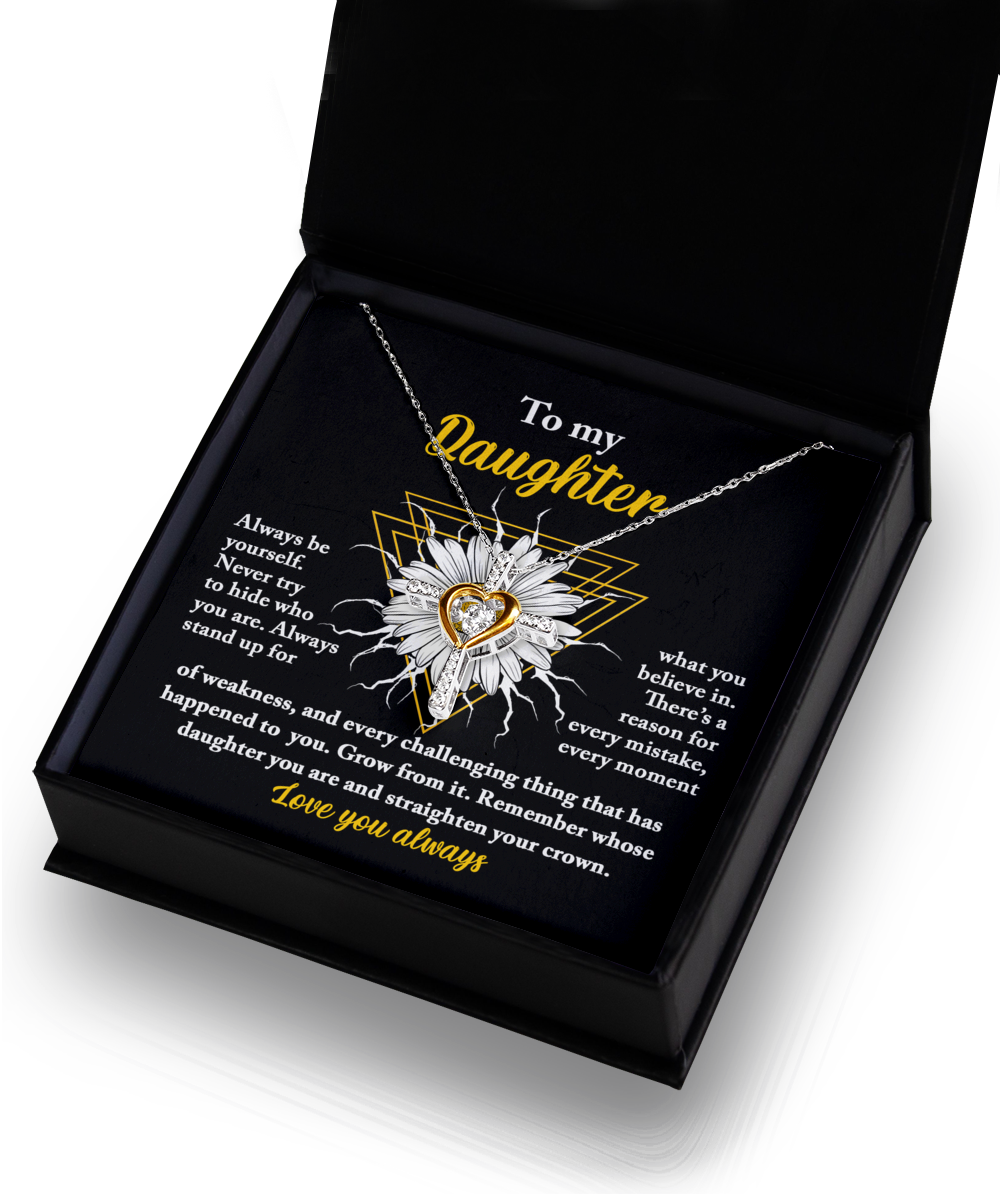 The "To Daughter-Be Yourself - Cross Dancing Necklace," featuring a sterling silver heart encasing a 14k gold-toned inner cross, is showcased in an open black gift box. The interior lid displays an encouraging message to a daughter in yellow and white text. A black insert within the box beautifully highlights the necklace.