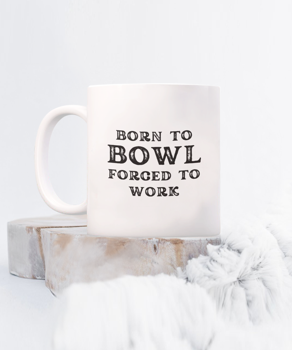 The "Born to Bowl Forced to Work" coffee mug features bold black text and is photographed on a wooden surface with soft fur accents. This unique item, not sold in stores, is printed and shipped from the USA as part of our best gift ideas for bowlers.