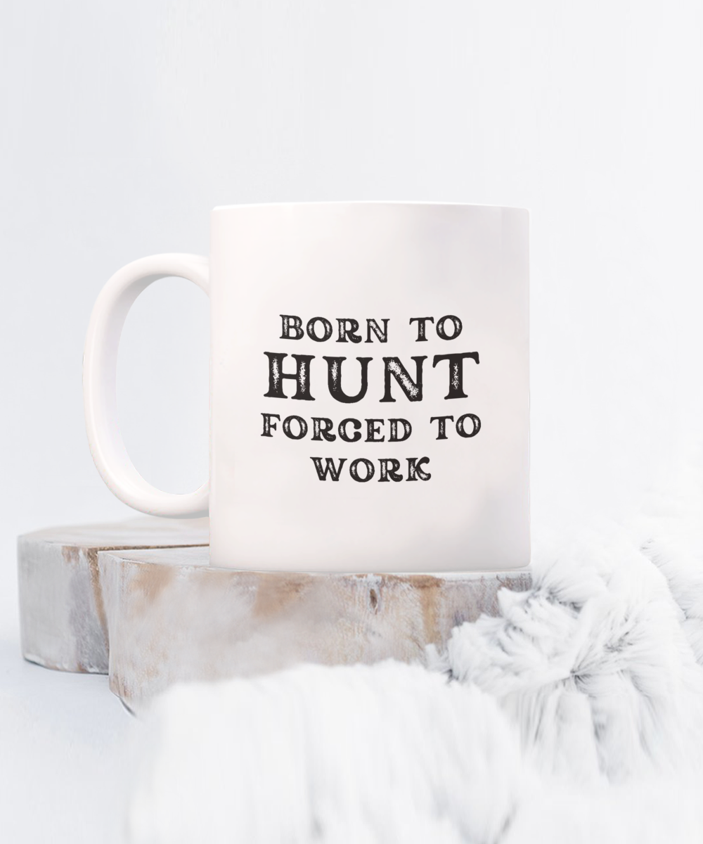 A white "Born to Hunt, Forced to Work" mug rests on a wooden surface beside soft, fur-like material. Printed and shipped from the USA for a limited time, this quality coffee mug ensures a guaranteed safe checkout. Ideal as a gift or appreciation cup for hunters.