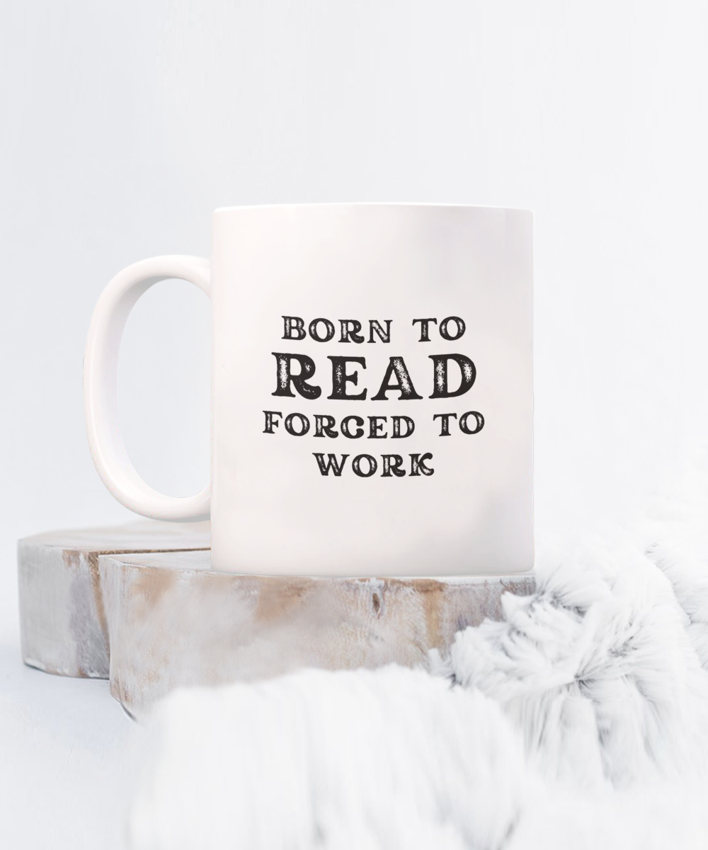 The Born to Read Forced to Work Coffee Mug, nestled on a wooden coaster with a fluffy white blanket nearby, is not available in stores, making it a unique find and perfect gift for book lovers.