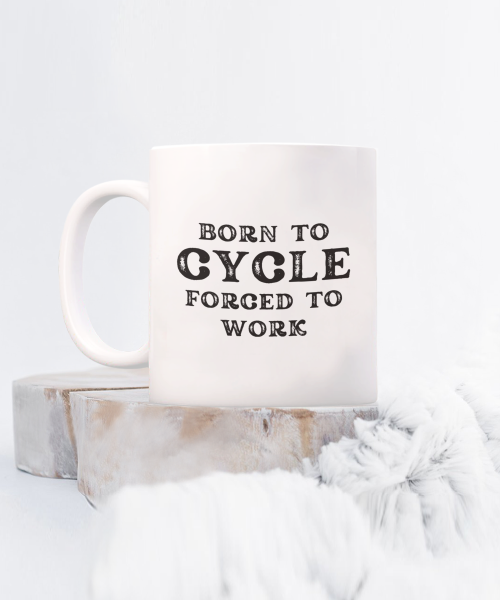 The "Born to Cycle, Forced to Work" coffee mug, featuring black text on a white surface, is displayed on wood against a fluffy white background. This USA-made design is exclusive and not available in stores.