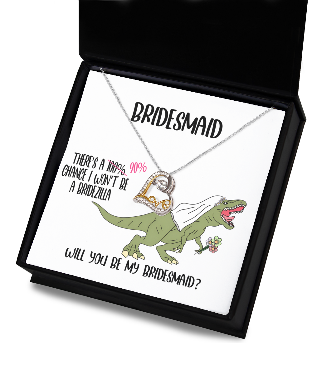 A Bridesmaid-Will You Be - Love Dancing Necklace is displayed on a card inside a black box. The card features a cartoon dinosaur in a veil holding flowers, with text asking, “Will you be my bridesmaid?”.