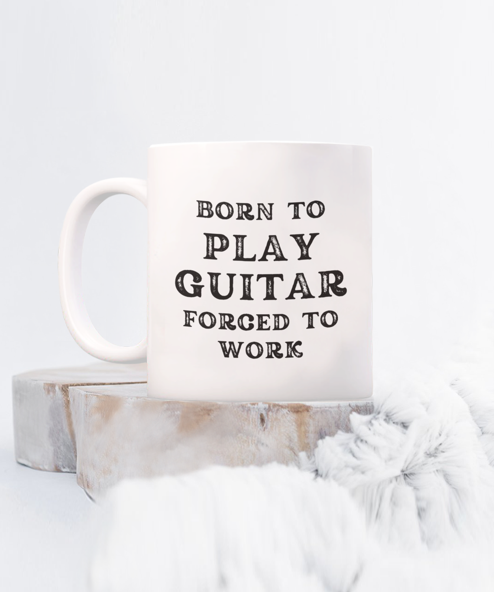 The "Born to Play Guitar, Forced to Work" coffee mug features black text on white, set on circular wooden coasters atop a fluffy light gray surface. Printed and shipped from the USA, this novelty item is available for a limited time. Perfect gift idea for guitarists!.