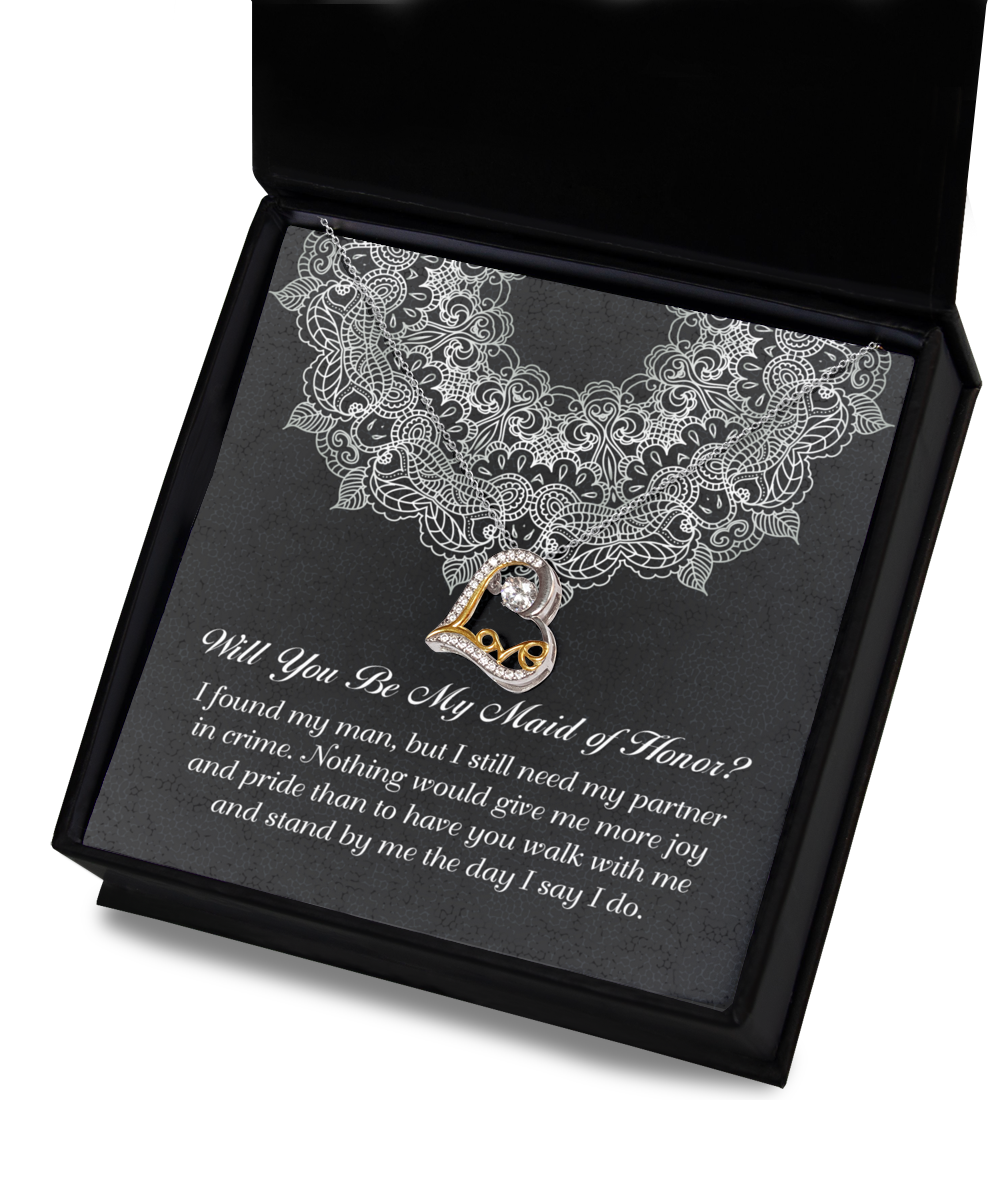 A black gift box with a silver lace design contains the "To Maid Of Honor-Partner In Crime - Love Dancing Necklace," featuring a heart-shaped pendant intertwined with the word "Love" and a gemstone. Inside, a note reads, "Will you be my Maid of Honor? I found my man, but I still need my partner in crime...