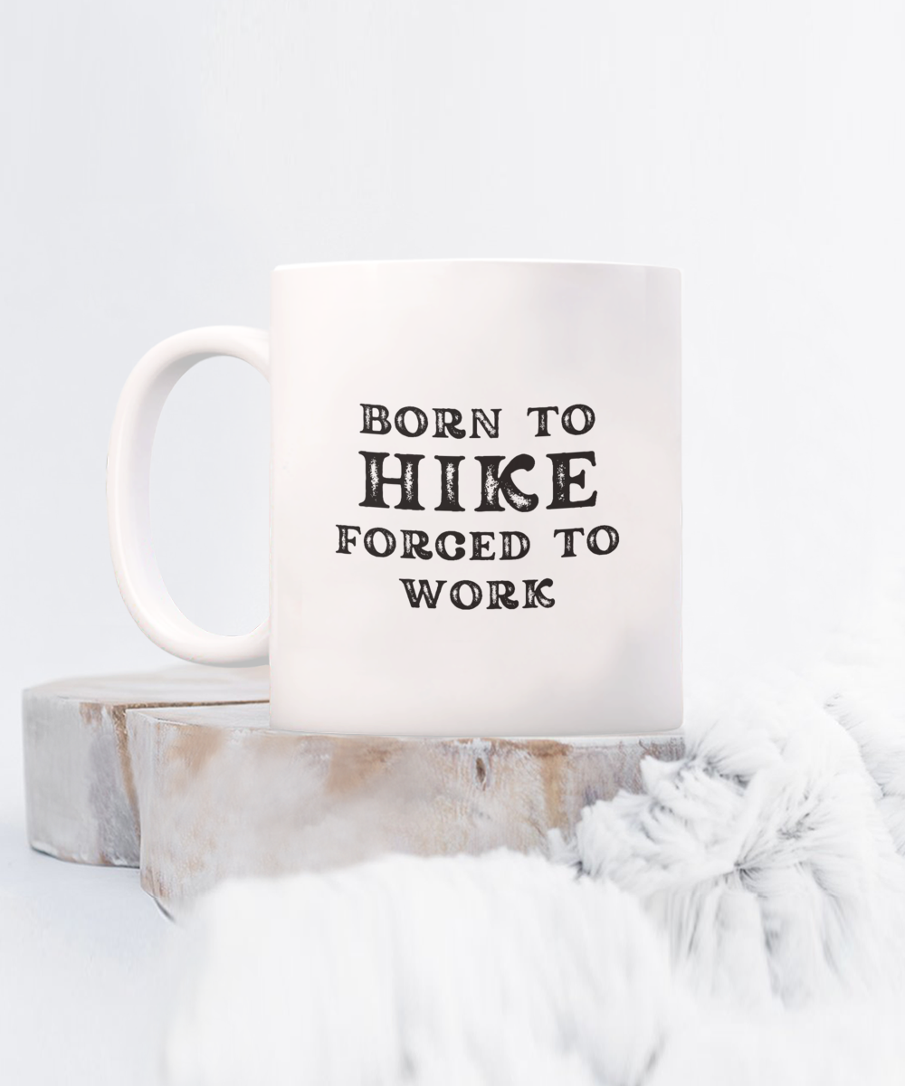 The "Born to Hike, Forced to Work" coffee mug sits on a wooden platform with a soft gray blanket in the foreground. This exclusive design isn't available in stores and is printed and shipped from the USA for a limited time, making it the best gift idea for hikers.