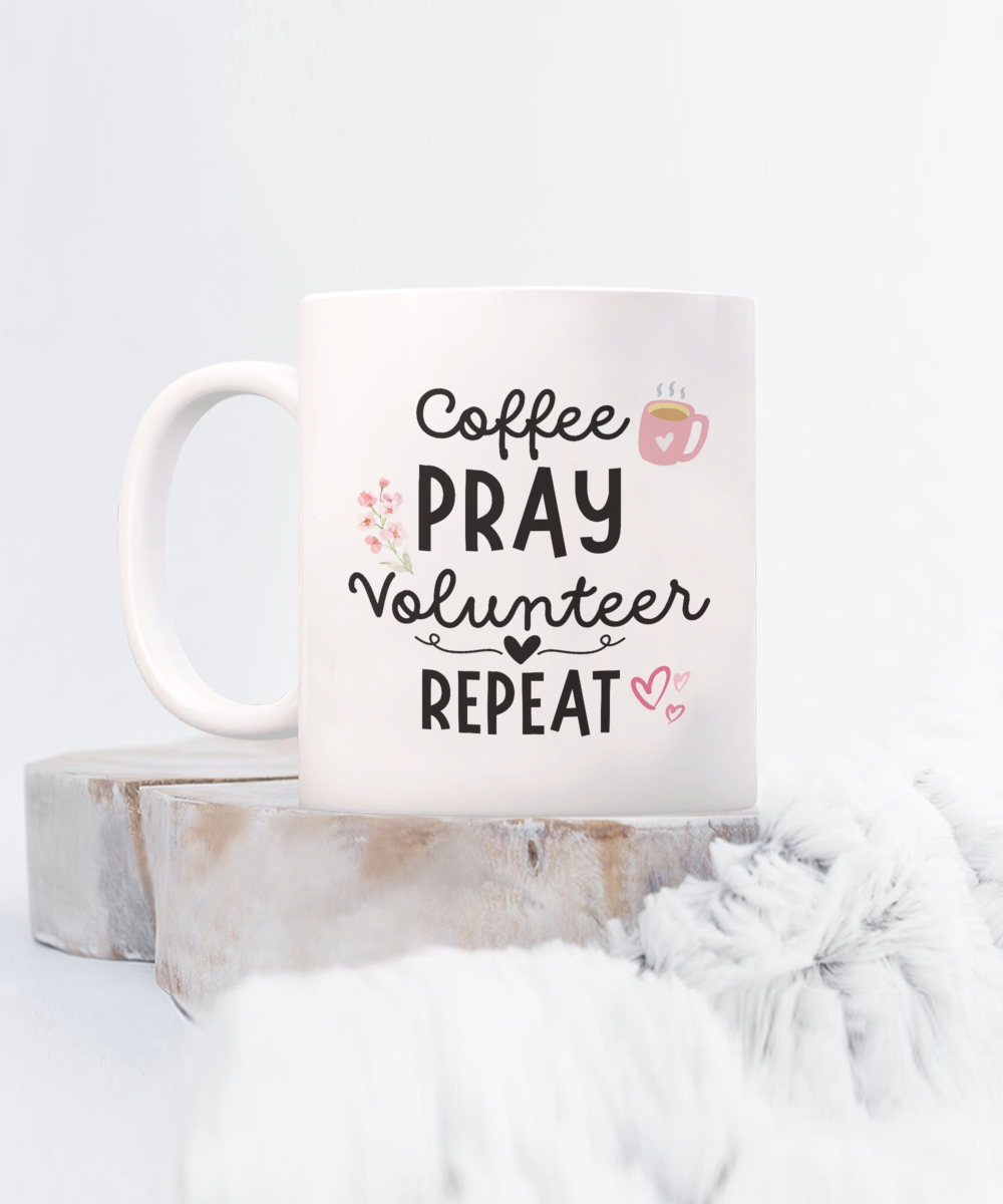 The Coffee Pray Volunteer Repeat Coffee Mug, featuring black text with heart and flower illustrations, sits on a wooden surface against a soft background. Perfect for limited-time gifting, this appreciation cup for volunteers is conveniently printed and shipped.