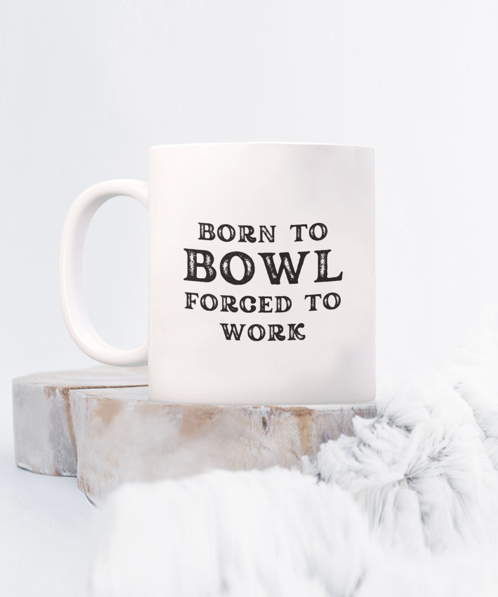 The "Born to Bowl Forced to Work" Coffee Mug rests on wood with soft fabric foreground. Not available in stores and printed in the USA, it's a unique find for bowlers seeking special gift ideas and appreciation cups.