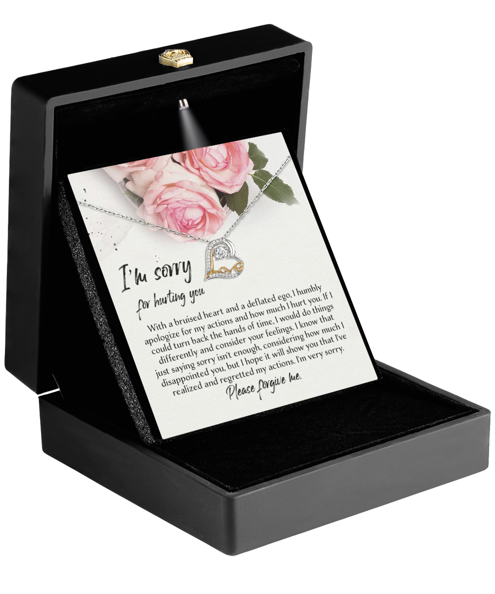 A black jewelry box is open to reveal the Sorry-Regretted My Actions - Love Dancing Necklace with two heart pendants, triple-plated in 14k gold. Inside the box is a note with an apology message, decorated with pink roses. The note reads, "I’m sorry for hurting you," followed by a heartfelt apology.