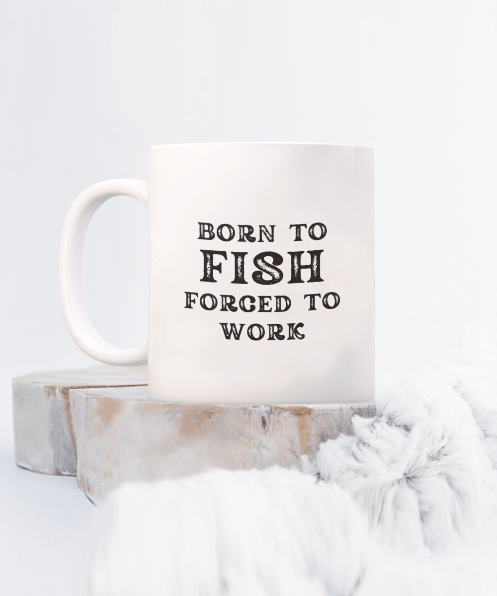 White mug with the phrase "Born to Fish, Forced to Work" rests on a wooden surface with soft fabric in view. Available for a limited time, this charming coffee mug is the perfect gift idea for anglers and fishermen, printed and shipped from the USA.