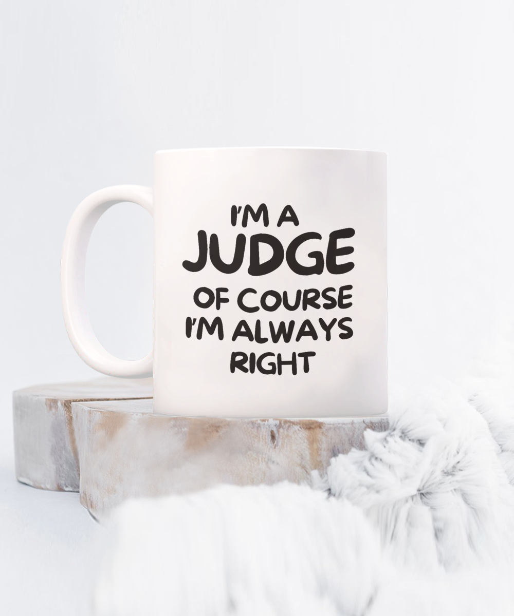 A white mug reads "I'm a JUDGE, OF COURSE I'M ALWAYS RIGHT" in bold black letters, placed on wood with gray fabric nearby. This unique design from the USA is available for a limited time. Product: I'm A Judge Of Course I'm Always Right Coffee Mug | Best Gift Ideas And Appreciation Cup For Judge.