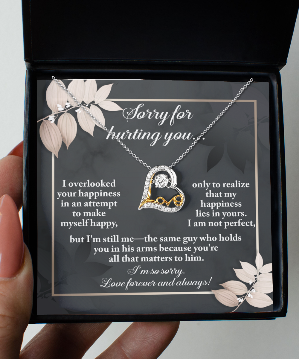 A heartfelt note within a black box accompanies the Sorry-Lies In Yours - Love Dancing Necklace, featuring a 14k gold loop enclosing a heart and a message apologizing for hurting the recipient while expressing enduring love and regret.