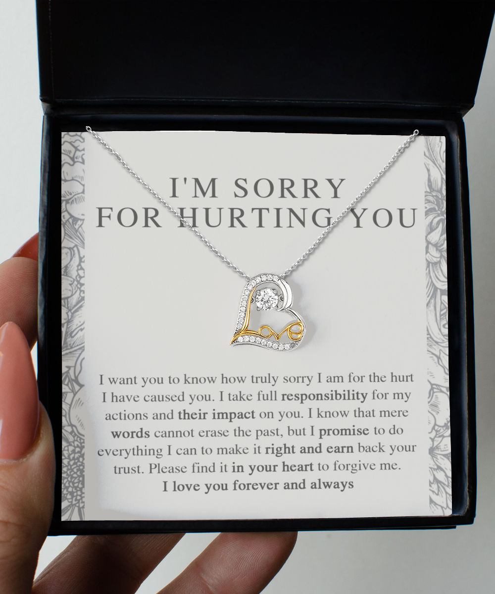 A hand holding an open black box containing a Sorry-Do Everything - Love Dancing Necklace with a heart-shaped pendant. The pendant, crafted in 14k gold and adorned with small gemstones, features the word "Love". Behind the necklace, a heartfelt apology message is printed, expressing regret and asking for forgiveness.