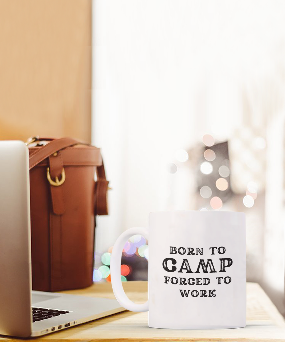 The "Born to Camp, Forced to Work" Coffee Mug, a limited-time-only piece printed and shipped from the USA, sits on a wooden table next to a laptop and brown leather bag.