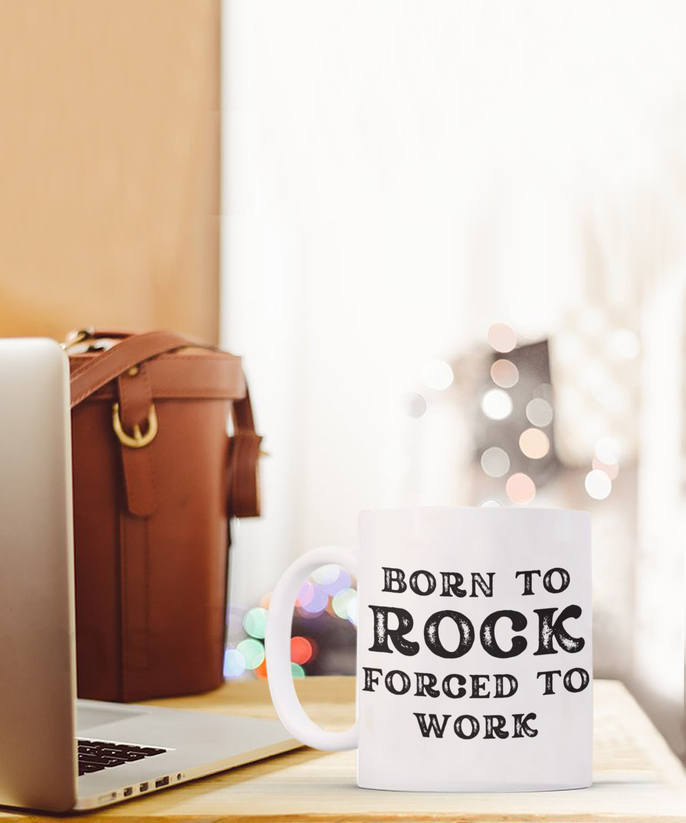 The "Born to Rock Forced to Work" coffee mug, perfect for adding personality to your workspace, sits by a laptop and brown leather bag. Printed and shipped from the USA, it's available for a limited time as an ideal gift or appreciation cup for rockers.