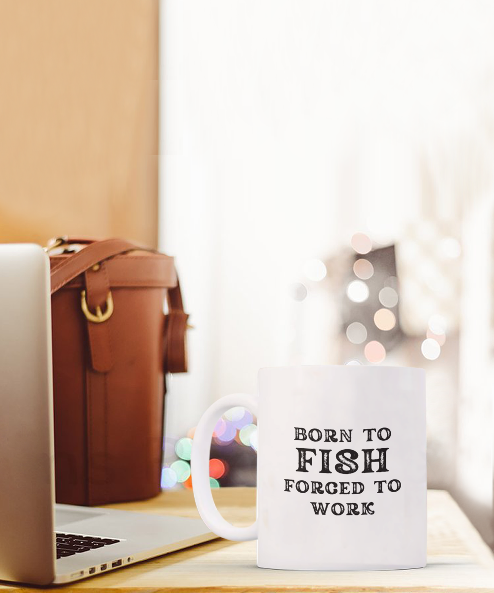 The "Born to Fish Forced to Work Coffee Mug," perfect for anglers and fishermen, sits on a table next to a laptop and leather bag. Printed and shipped from the USA, it's available for a limited time.