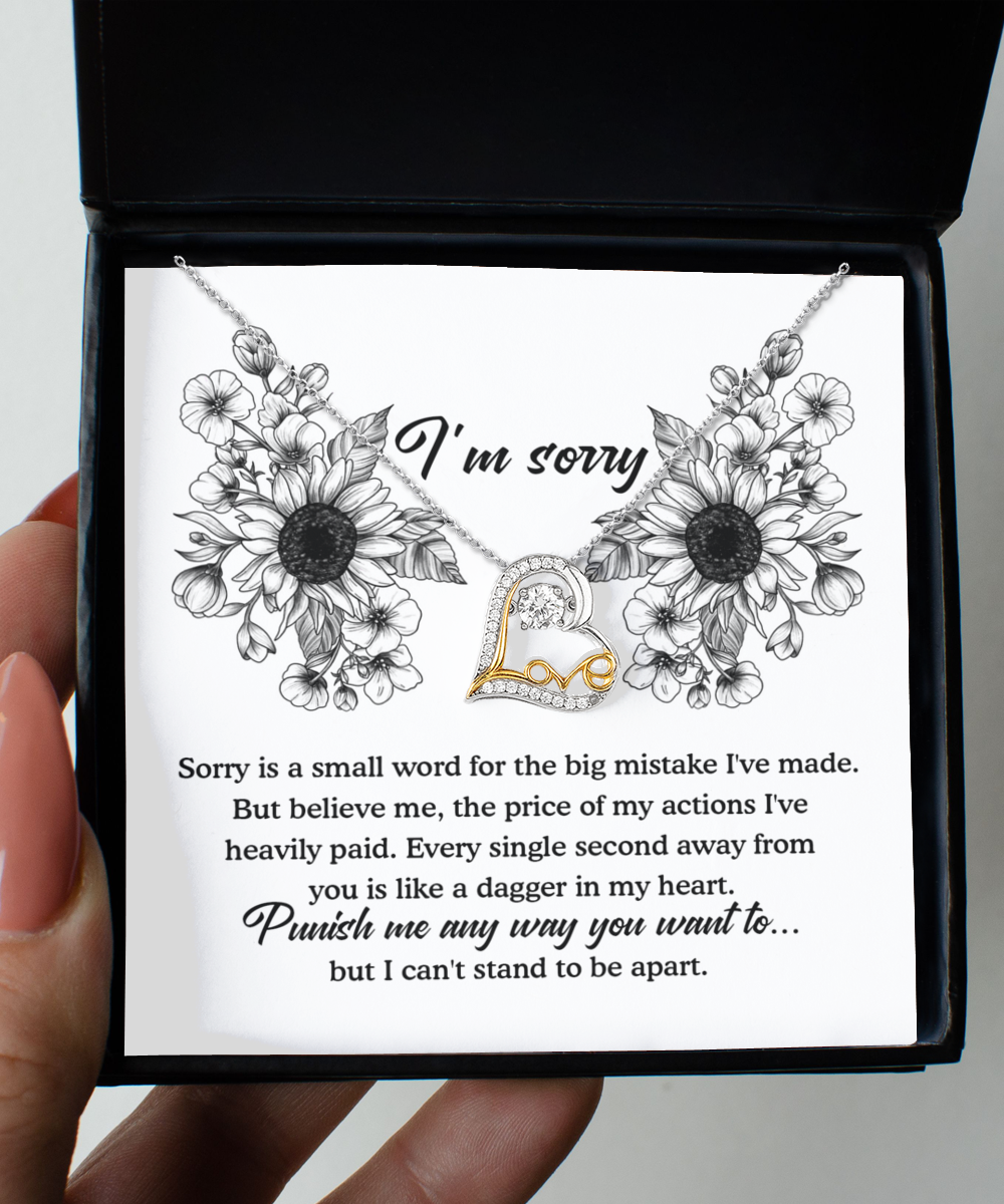 A person holds an open black gift box containing a Sorry-Small Word - Love Dancing Necklace, a sterling silver necklace with a "Love" pendant, and a card adorned with floral illustrations. The card reads, "I'm sorry. Punish me any way you want to… but I can’t stand to be apart," expressing an apology in the text.