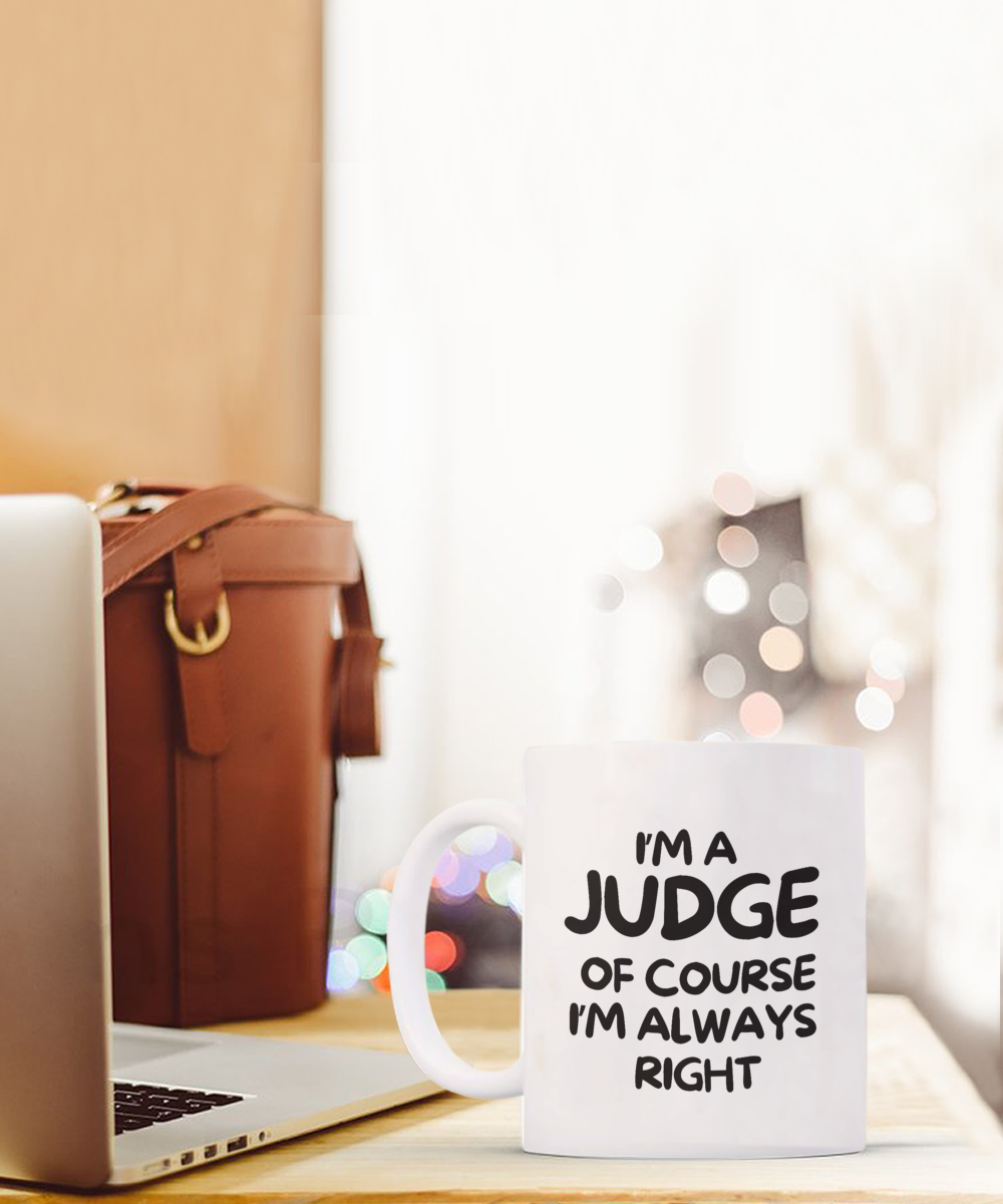 On a desk beside a laptop and a brown leather bag, there's an "I'm A Judge Of Course I’m Always Right Coffee Mug" available for a limited time. It's printed and shipped from the USA with guaranteed safe checkout.