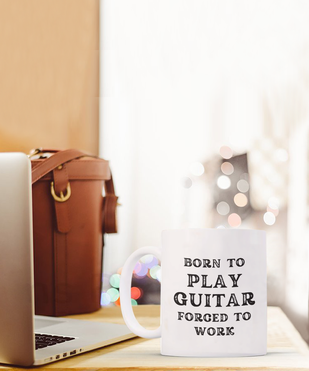 The "Born to Play Guitar Forced to Work" coffee mug is shown near a laptop and leather bag. Shipped from the USA, this distinctive mug is offered for a limited time.