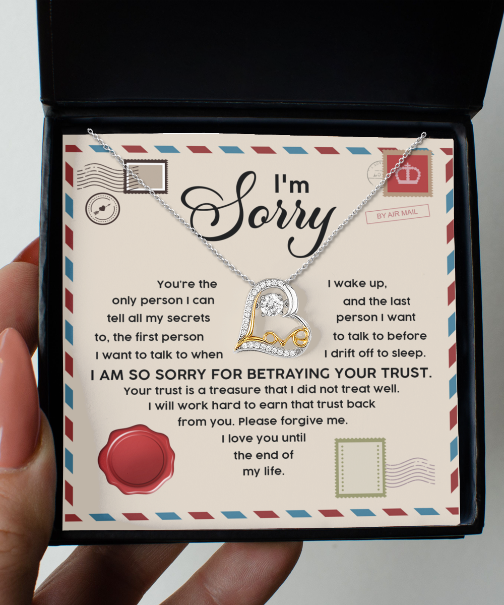 A person holds an open box containing a Sorry-The Only - Love Dancing Necklace, which features a sterling silver heart pendant. The box's lid has an apology message printed on it, including the phrase "I am so sorry for betraying your trust.