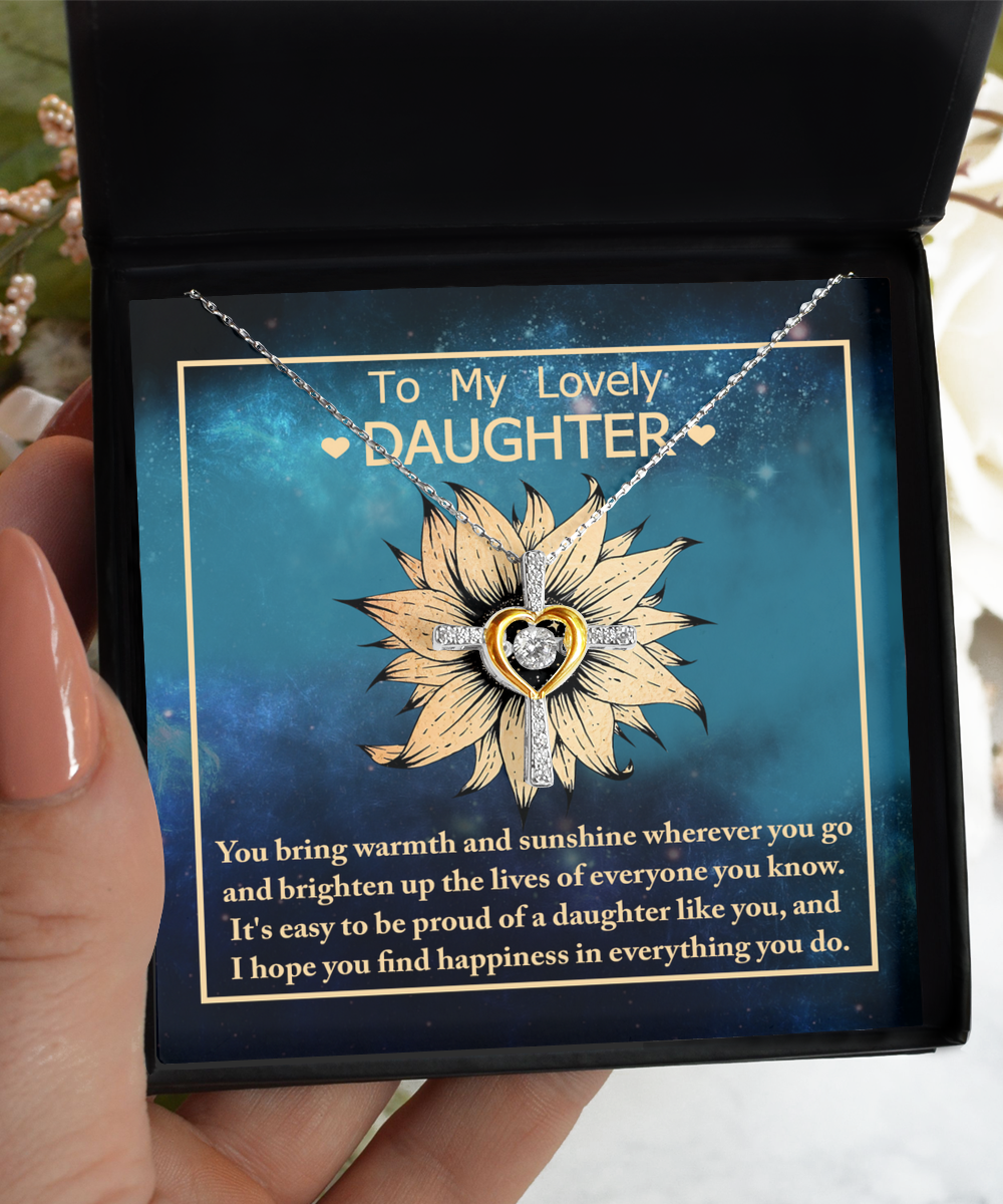 A hand is holding an open black jewelry box containing the "To Daughter-Be Proud Of - Cross Dancing Necklace". The necklace features a sunflower pendant with a heart-shaped center. The background inside the box displays a heartfelt message: "To My Lovely Daughter...I hope you find happiness in everything you do.