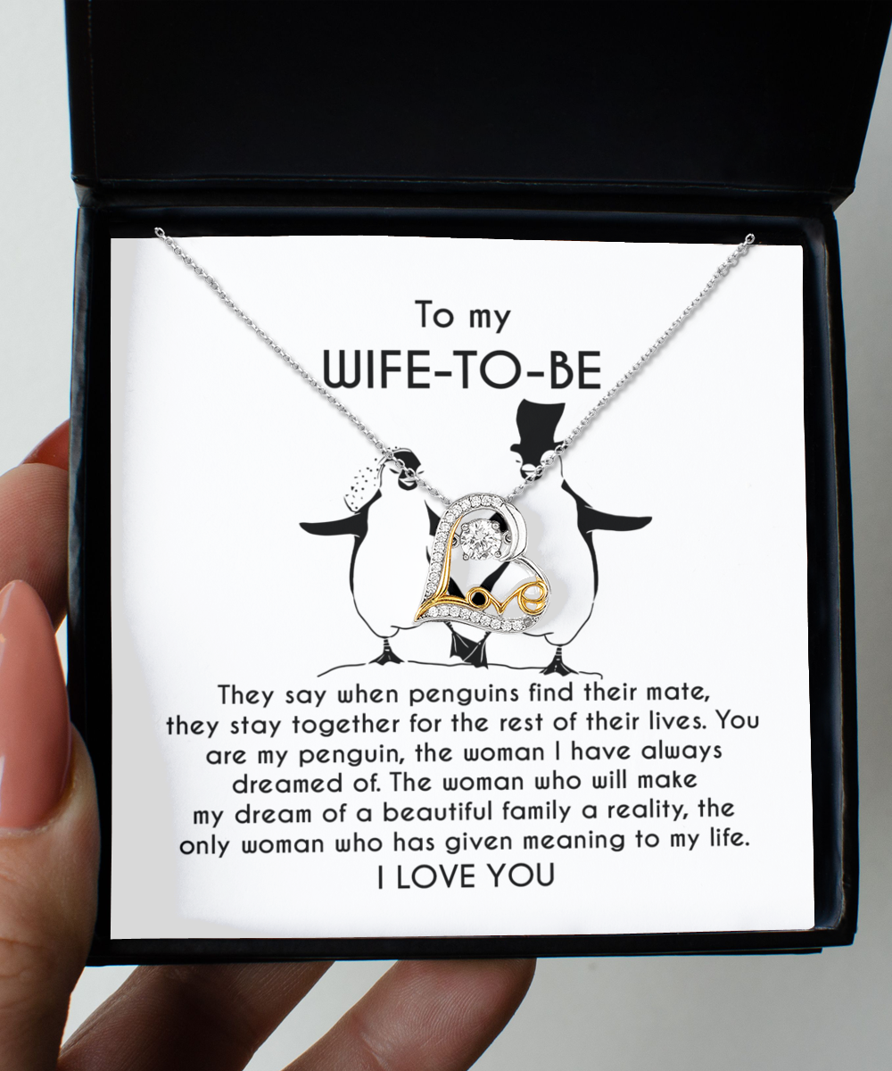 A silver heart-shaped necklace with an intertwined 14k gold plated heart centerpiece sits on a message card inside a black box. The card reads: "To my WIFE-TO-BE, I LOVE YOU" with a monologue about love and penguins. This is the Wife-to-be-My Penguin - Love Dancing Necklace.