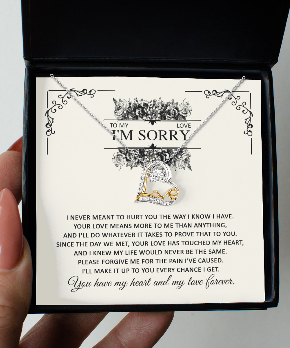 A hand holds a box containing the Sorry-Every Chance - Love Dancing Necklace, a sterling silver hypoallergenic piece with a "Love" pendant. The box lid has a heartfelt apology note adorned with floral designs, expressing regret and a commitment to make amends. Both the pendant and note symbolize deep affection and sincere apology.