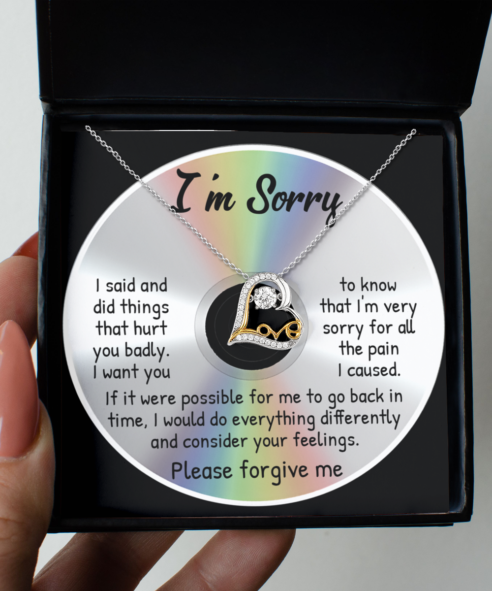 A hand holds an open box containing a Sorry-Back In Time - Love Dancing Necklace with a heart pendant, placed on a CD with an apology message printed on it.