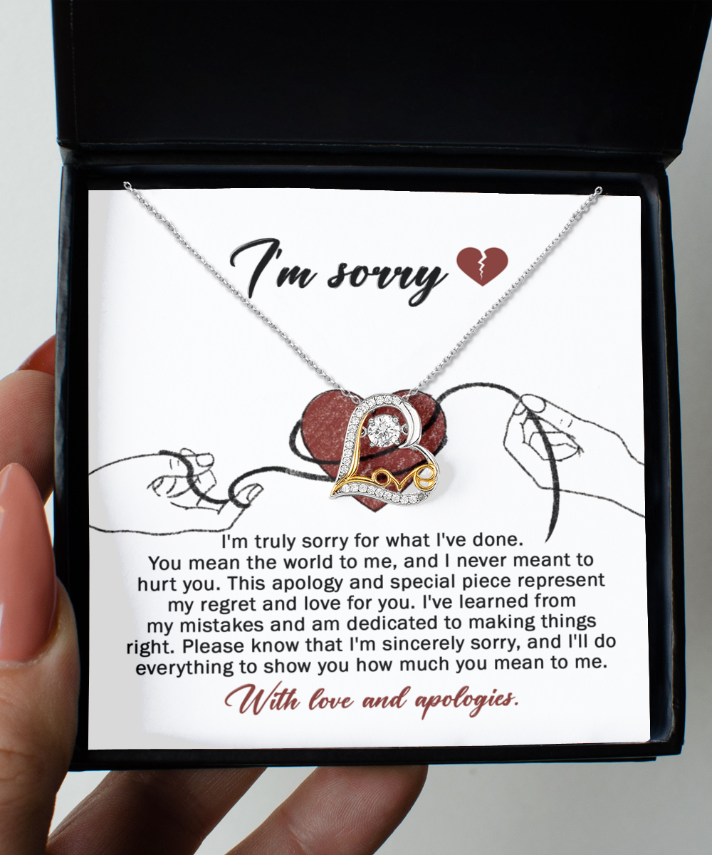 The "Sorry-Mean The World - Love Dancing Necklace," featuring a heart pendant, is displayed in a black box. The interior of the box is lined with 14k gold and includes an apology message expressing regret and love, along with an illustration of two hands holding red string.