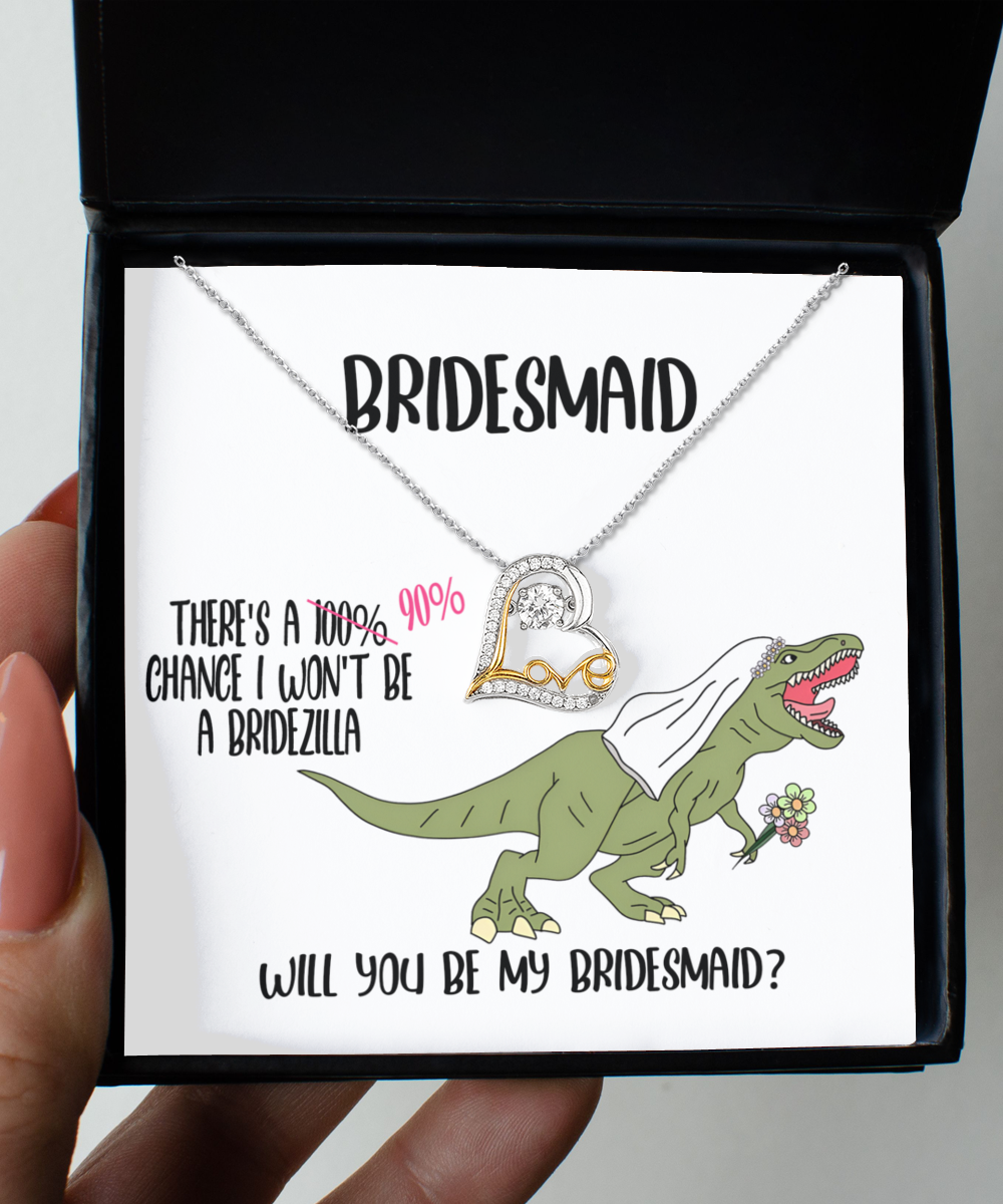 A Bridesmaid-Will You Be - Love Dancing Necklace is displayed on a card inside a black box. The card features a cartoon dinosaur in a veil holding flowers, with text asking, “Will you be my bridesmaid?”.